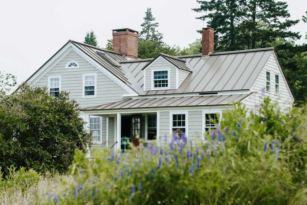 A modern, tan metal roof covers a white-trimmed home surrounded by trees and shrubbery.